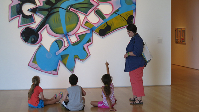 Three children sit on a floor an point at an artwork.