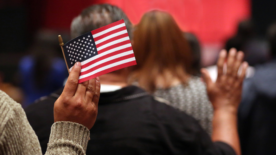 Back view of people taking the citizenship oath with their right hands raised. One person is holding a U.S. flag in his raised hand.