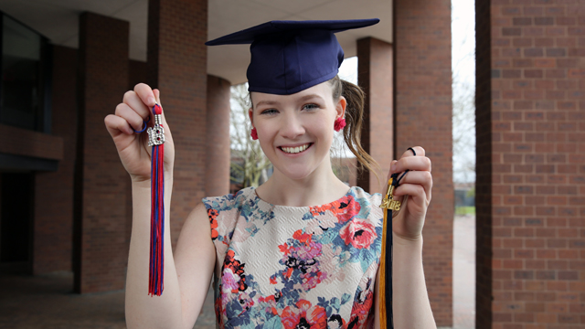 Grace Palcic wearing a graduation cap and holding tassels from two different schools