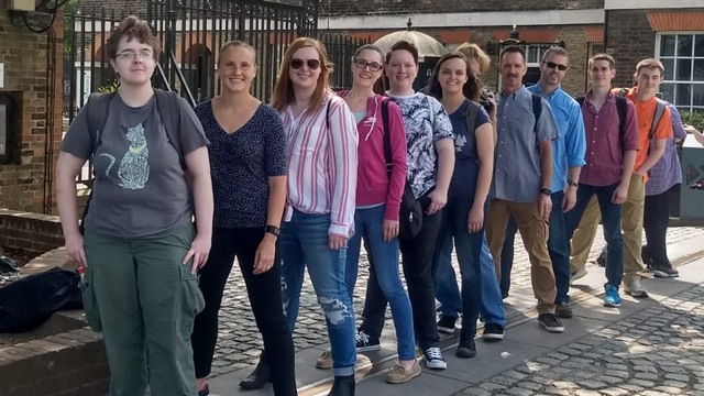 Alt text: Eleven people straddle a metal bar on the ground in Greenwich, England. The bar represents the location of the prime meridian, the imaginary line of zero longitude around the earth from the North to South Poles.