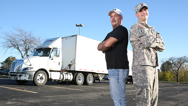 Two veteran CDL students pose back to back in front of a long, white semitruck.