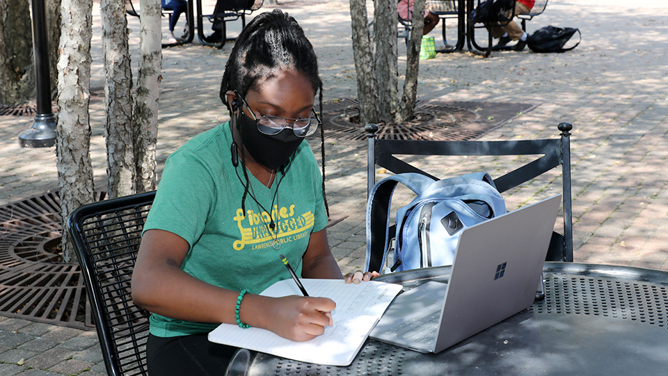 Female student, wearing a mask, studying with her laptop at an outdoor table