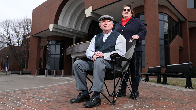 Steve Adams is seated in a folding chair in front of the Midwest Trust Center. His wife, Lisa, stands behind him.