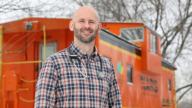 Thomas Bristow, wearing a stethoscope and standing in  front of the BNSF caboose on campus