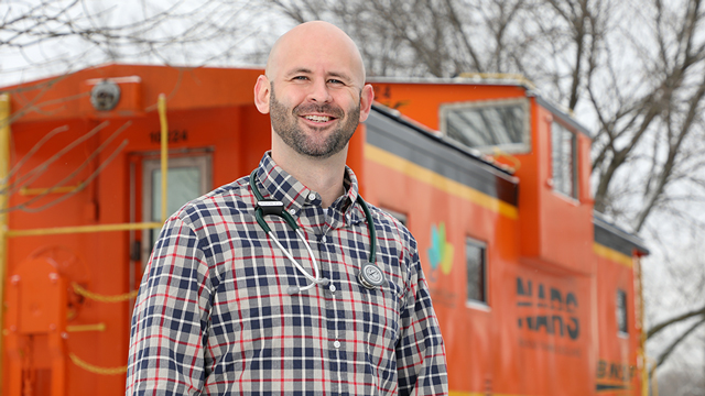 Thomas Bristow, wearing a stethoscope and standing in front of the BNSF caboose on campus