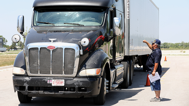 Photo of student behind wheel of semi-truck in campus parking lot, receiving instruction from trainer standing next to truck, through truck window.