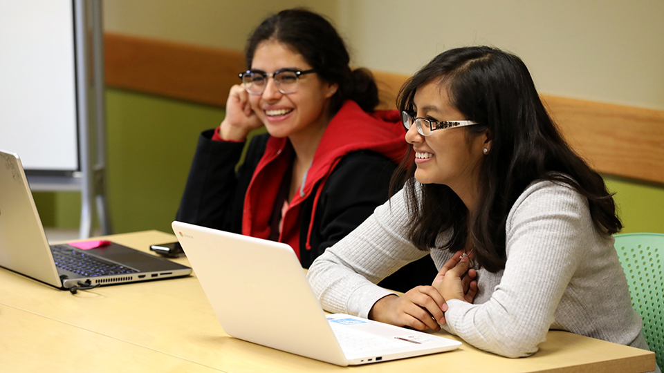 Two smiling female students with their laptops in a classroom