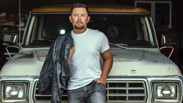 Scotty McCreery holding a denim jacket over his shoulder and posing in front of a vintage Ford truck