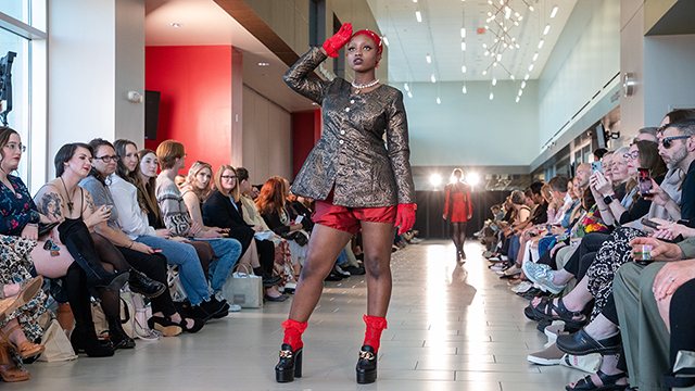 An audience is watching a fashion show. The model in front, photographed from a low angle, wears a tight red pearled cap, a pearl choker with pendant, black and gold brocade buttoned jacket, red poofy shorts with elastic cuffs, red lace gloves and socks, and high black mule heels.