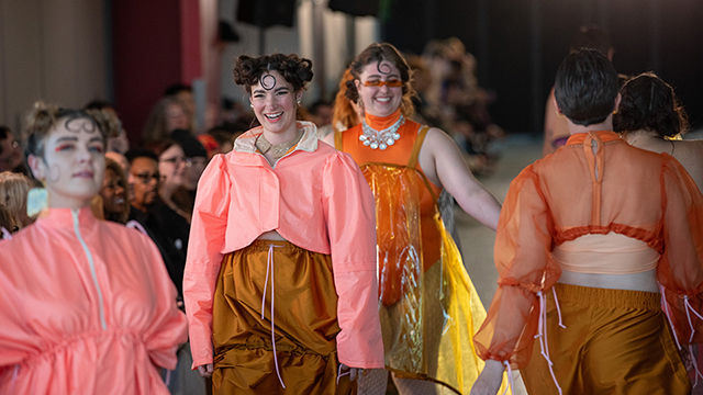 A group of models strut the runway, wearing various outfits in shades of coral, brown, gold and orange.