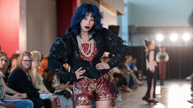 : A woman with dark blue-black hair and deep red lipstick strikes a pose on the runway. She is wearing a puff-sleeved black jacket, white pearls, a dark red shirt with gold dot pattern, dark red-gold shorts, and black heels with low socks.