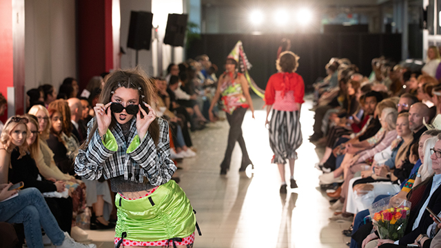 A crowd sits on two sides of the runway as models walk on it. At the front of the runway is a tattooed woman wearing a lime green satin skirt with pink and red polka dot accents, a black and white plaid jacket with lime green satin lining, and sunglasses.