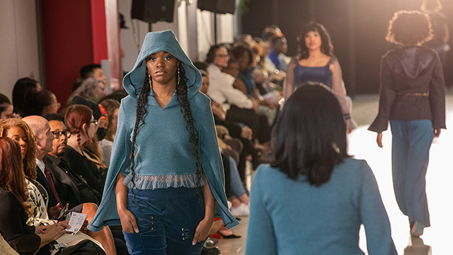 : A line of models walks down the runway in two directions as an audience looks on.