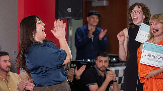 A woman with long dark hair wearing a black shirt and brown and gold skirt smiles excitedly while clapping her hands together. An audience applauds.