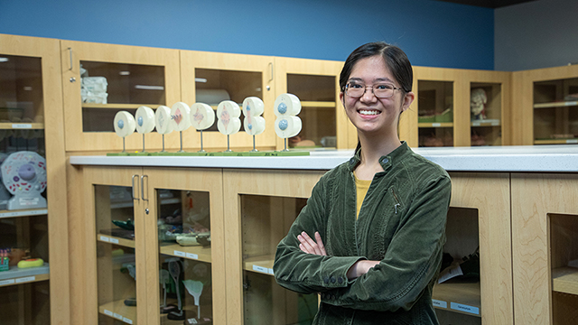 Biology tutor Kaitlyn Sy stands in a science lab room with cabinets containing scientific teaching tools around her. To her right is a display showing how a cell divides.  