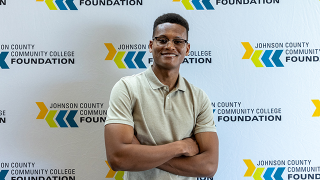 Shayne McCrae wears glasses and a tan polo shirt. He stands with arms crossed in front of a large backdrop that reads “Johnson County Community College Foundation” multiple times.