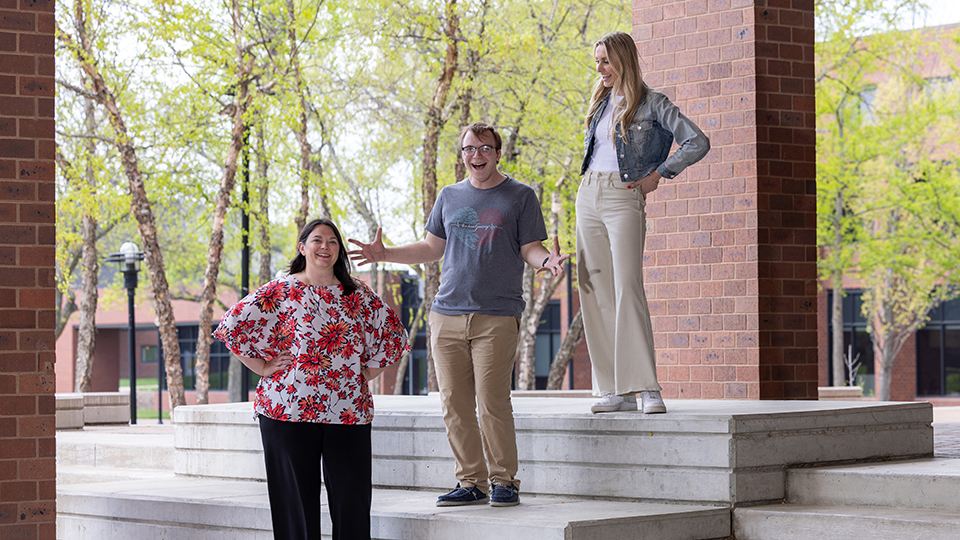 A group of three people stand on large stairs leading up to the right: A woman stands to the left on the bottom step with her hands on her hips, a man with his arms out to the side in the middle, and another woman stands on the top step looking down at her companions.
