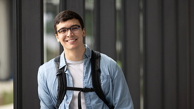 Isaac Zarate smiles while wearing glasses, a white t-shirt, a blue button-down shirt, and the straps of a backpack. He stands in front of a nondescript architectural background.