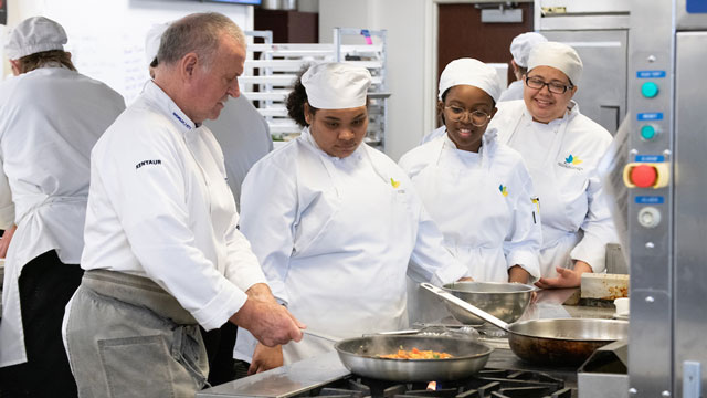 Guest chef Rick Stephen, on the left, cooks a pan of food over a gas stove while three JCCC Chef Apprenticeship students dressed in white aprons and hats look on in a JCCC Culinary kitchen classroom.