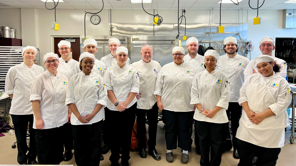 Thirteen JCCC Chef Apprenticeship students (seven on the left and six on the right) pose with guest chef Rick Stephen (center) for a group photo in a JCCC Culinary kitchen classroom.