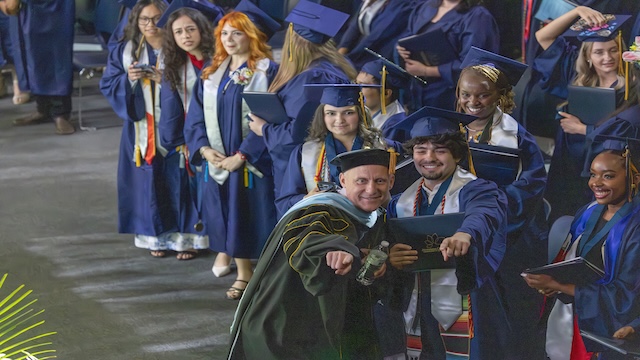 Dr. Miksa poses with a male student at graduation. The student proudly holds his diploma. Both point toward the camera. Many other students in their regalia surround them, holding their diplomas and smiling.