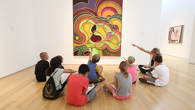 A group of young children sitting in a semi-circle on the floor in front of a large, colorful painting in the Nerman during a class