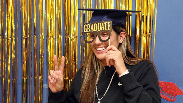 A smiling JCCC Grad stands in front of a festive background while holding a prop glasses that say graduate over her face