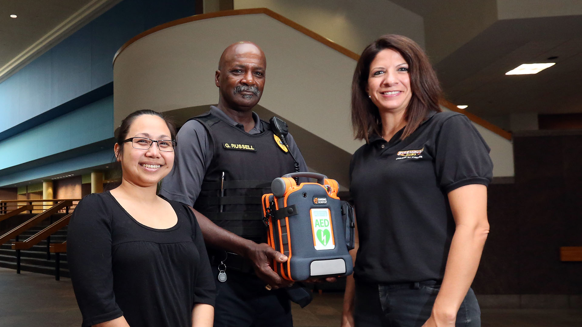 Em Smail, supervisor of JCCC Dining Services, Gregory Russell, chief of police, and Tara Ruff, franchise owner of the Firehouse Subs in the JCCC food court, show off the new campus AED device. Photo by Susan McSpadden/JCCC