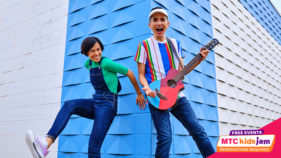 Christina and Andres stand in front of a blue and white wall. Christina is smiling and leaning against the wall. Andres is singing while holding a colorful guitar.