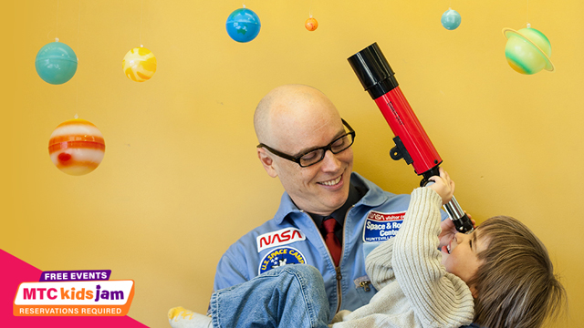 A smiling Dino O'Dell looks down at a young boy who is using a telescope. Dino is wearing a blue jacket covered in NASA patches.