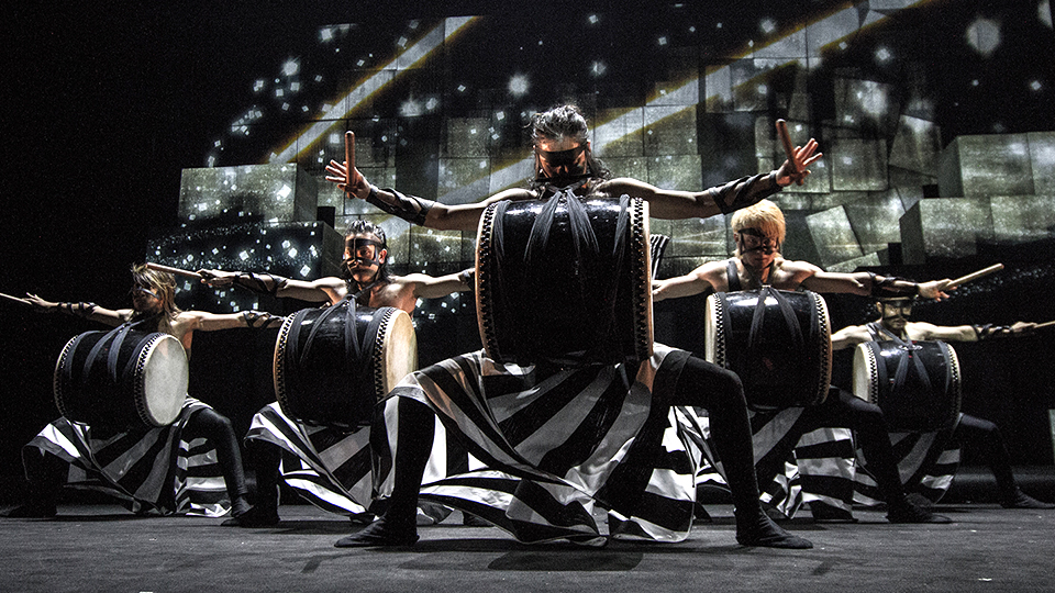 Five members of Drum TAO perform on stage. They stand wearing black and white striped costumes with arms outstretched and in front of a screen displaying dynamic graphics.