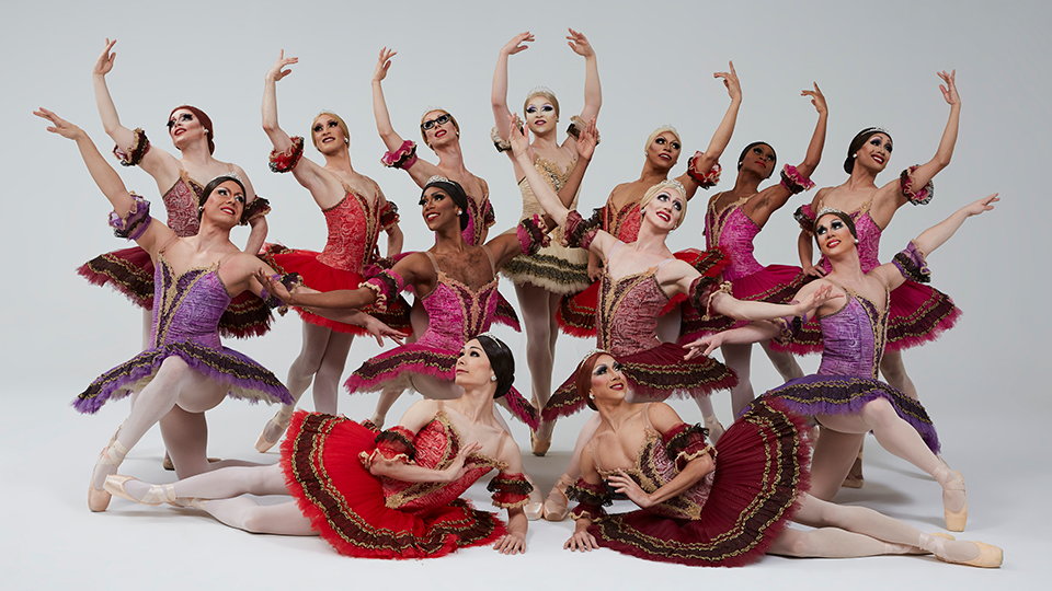 Thirteen members of Les Ballets Trockadero de Monte Carlo pose gracefully as a group with arms outstretched and toes pointed.