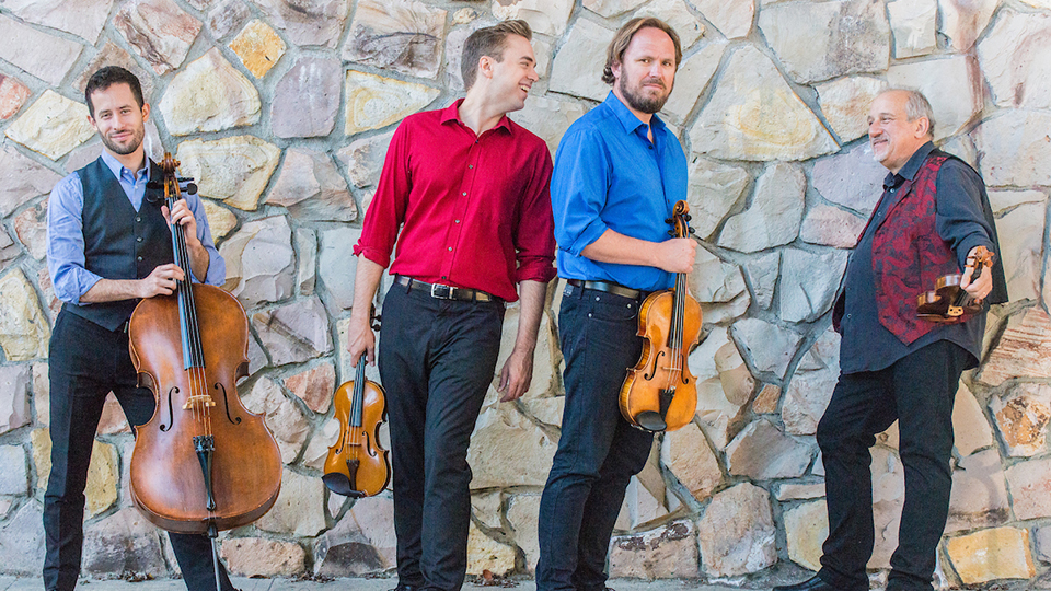 The four members of Quartet San Francisco stand in front of a stone wall while smiling and holding their instruments