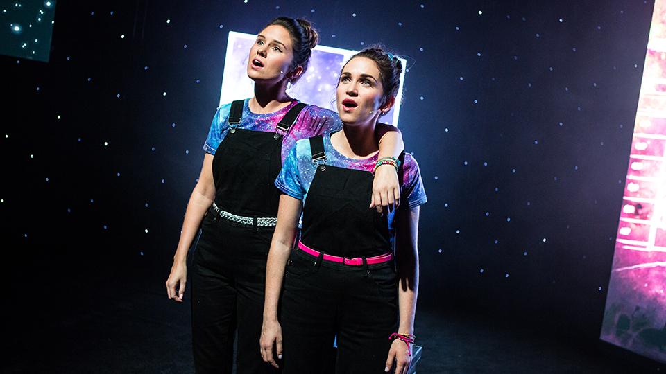 Two young women sing together in front of a space-themed backdrop.