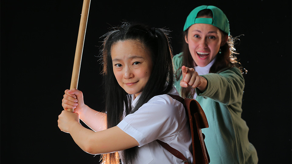 Two actresses on stage - one is holding a baseball bat and the other is pointing at her