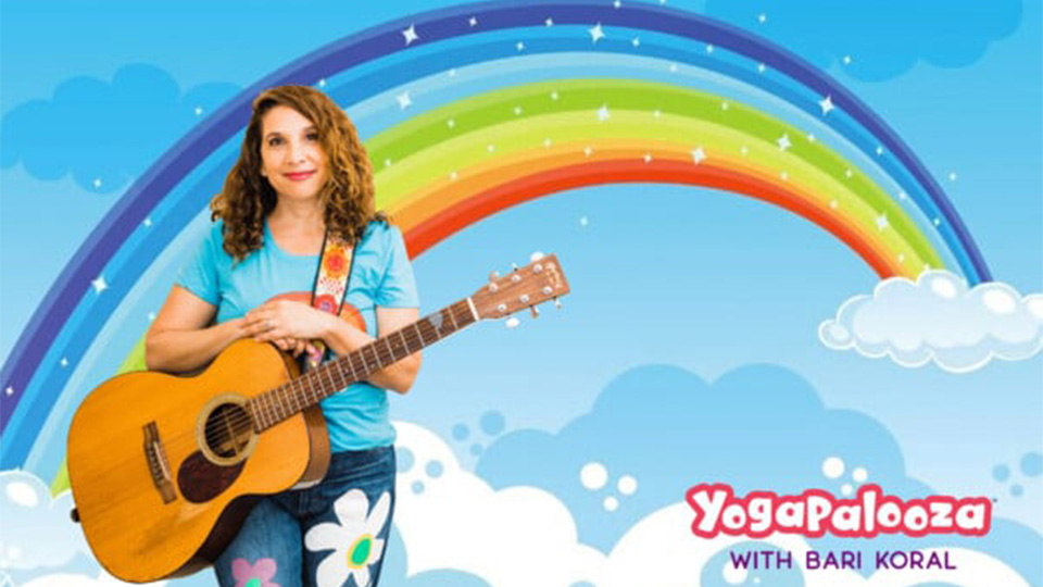 Bari Koral standing in front of a rainbow with her guitar and the words yogapalooza