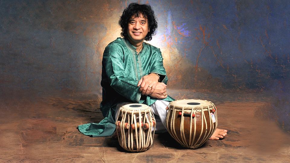 Zakir Hussain smiles at the camera while sitting comfortably on the floor behind two drums and in front of a backdrop painted in abstract colors of blues and tans