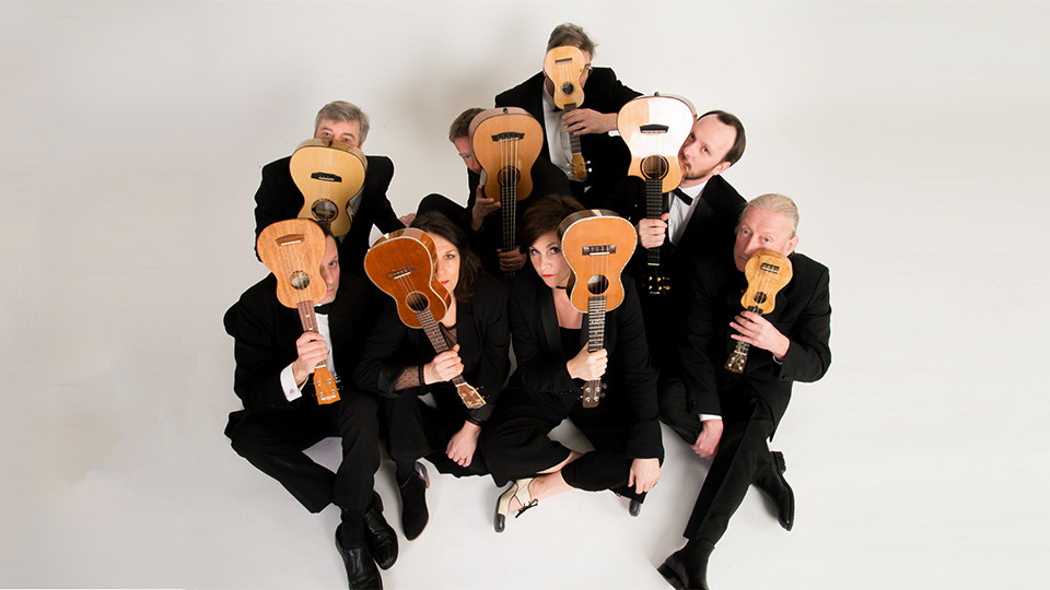 the 8 members of the ukulele orchestra sitting cross-legged on the floor and holding their ukuleles in front of their faces