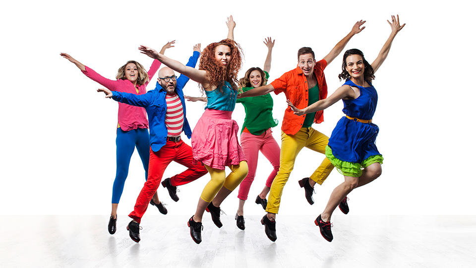 six dancers wearing brightly colored clothes jump to the side with their hands raised in the air