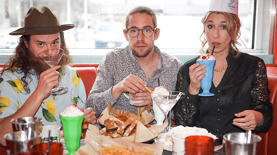 three people sitting in a diner booth drinking and eating