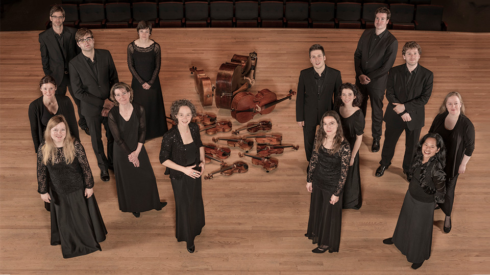 The 14 members of Les Violons pose for a picture. The picture shows them from over head standing in two groups on stage with their violins arranged on the floor in the middle of the stage.