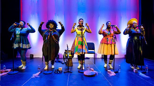 five singers in colorful outfits stand on stage with their hands raised
