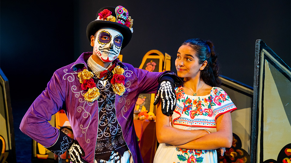 A man in traditional Day of the Dead skull makeup stands next to a woman wearing a traditional Mexican dress