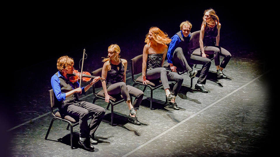 five members of the StepCrew sitting on stage in chairs - one of them is playing a fiddle
