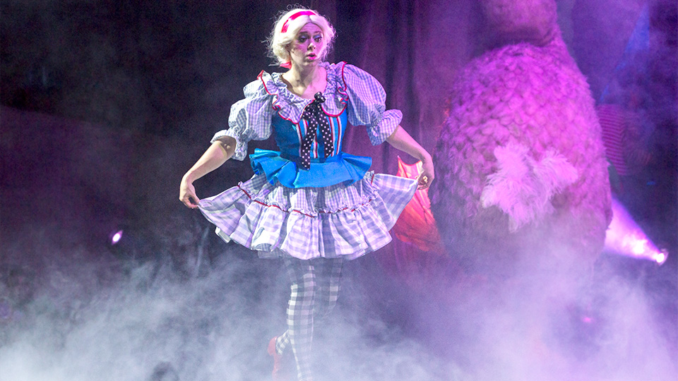 An actress portraying Alice walks through fog on stage while holding her skirt out