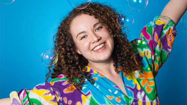 Kayla Sandusky is smiling with her arms raised and standing in front of a bright blue background with soap bubbles around her 
