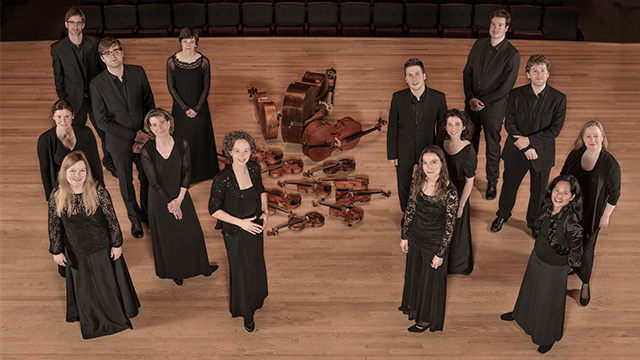 The 14 members of Les Violons pose for a picture. The picture shows them from over head standing in two groups on stage with their violins arranged on the floor in the middle of the stage.