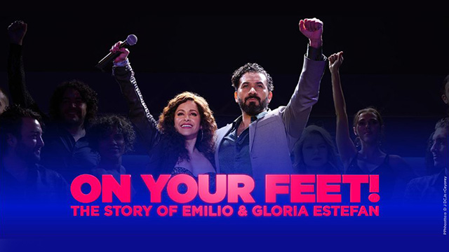 two actors portraying Gloria and Emilio Estefan stand arm-in-arm on stage with their other arms raised while looking out toward the viewer. The words On Your Feet The Story of Emilio and Gloria Estefan are superimposed in front of them. 