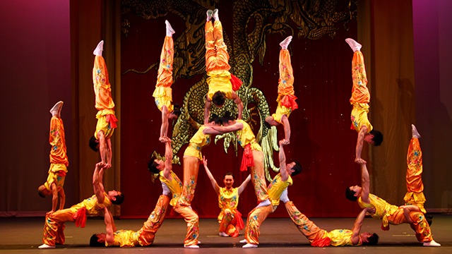 a group of 16 acrobats perform a stunt on stage where seven of them are doing hand-stands with the other acrobats as bases