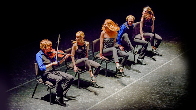 five members of the StepCrew sitting on stage in chairs - one of them is playing a fiddle